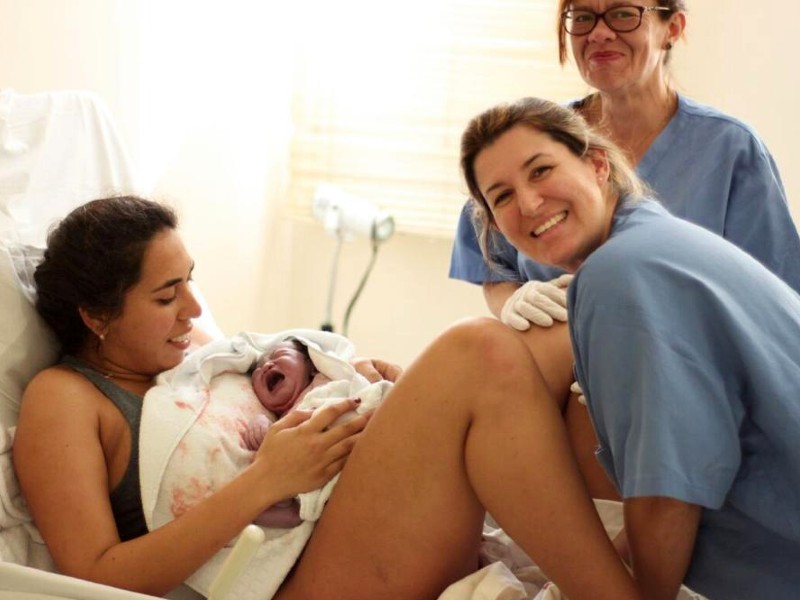 Foto de Thais Talarico com uma mãe que acabou de dar à luz. Thais está de uniforme azul, olhando para a frente e sorrindo. Na frente dela, a mãe segura a bebê recém-nascida embrulhada em uma toalha branca e sorri. Outra enfermeira aparece atrás das duas, também sorrindo